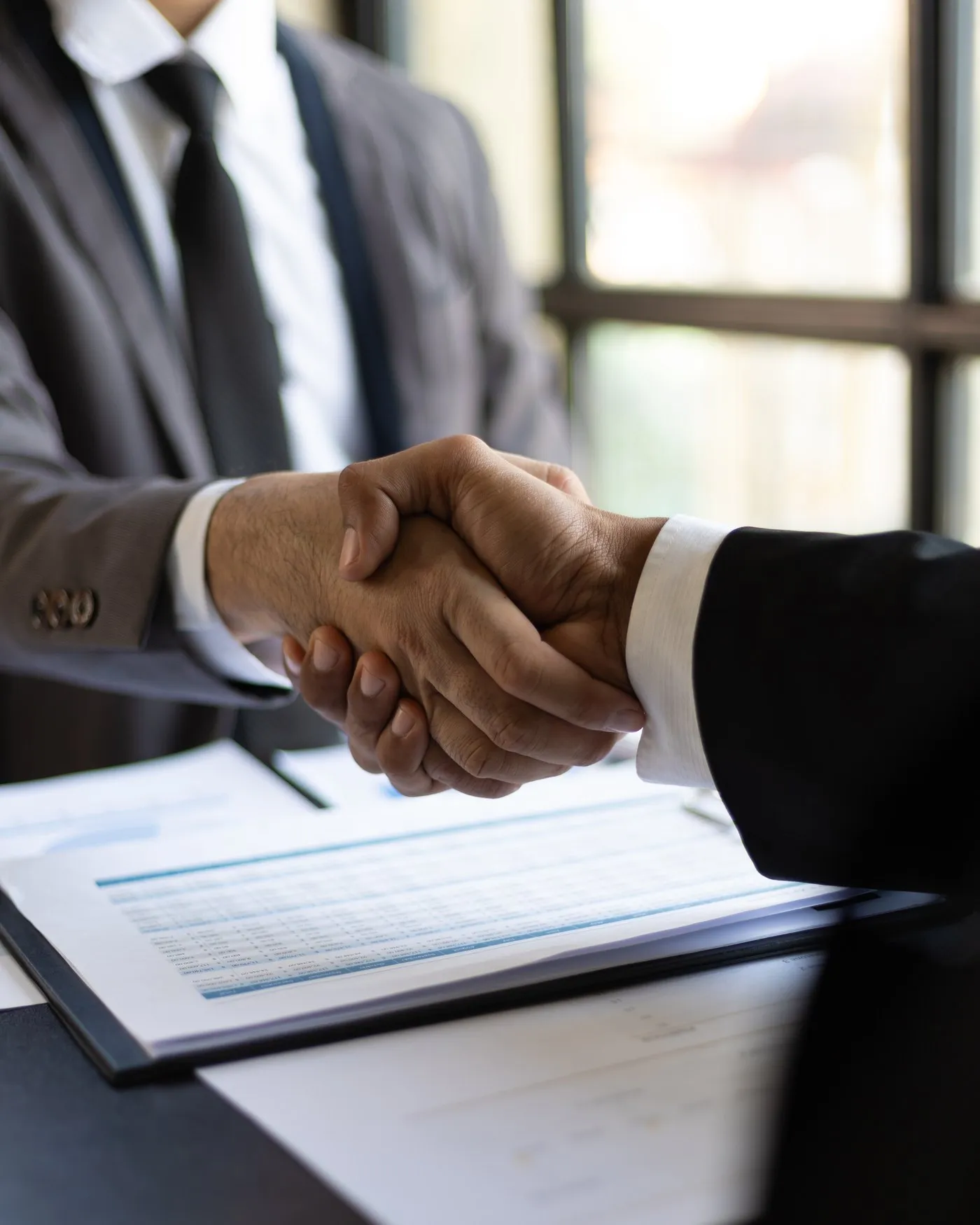 Two professionals shaking hands across a desk over financial paperwork